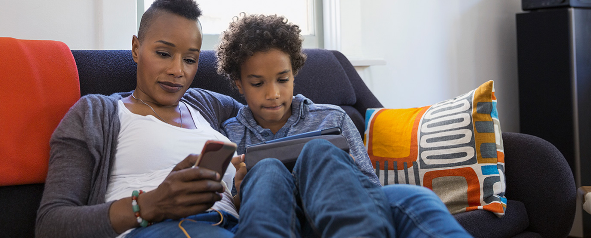 A mother and child sitting together on a couch, each using their own mobile device